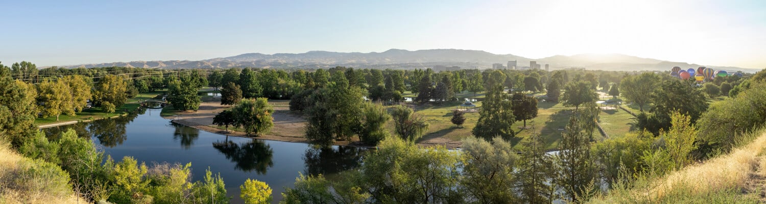 A scenic park with a calm lake, lush green trees, and mountains in the background under a clear sky in boise id