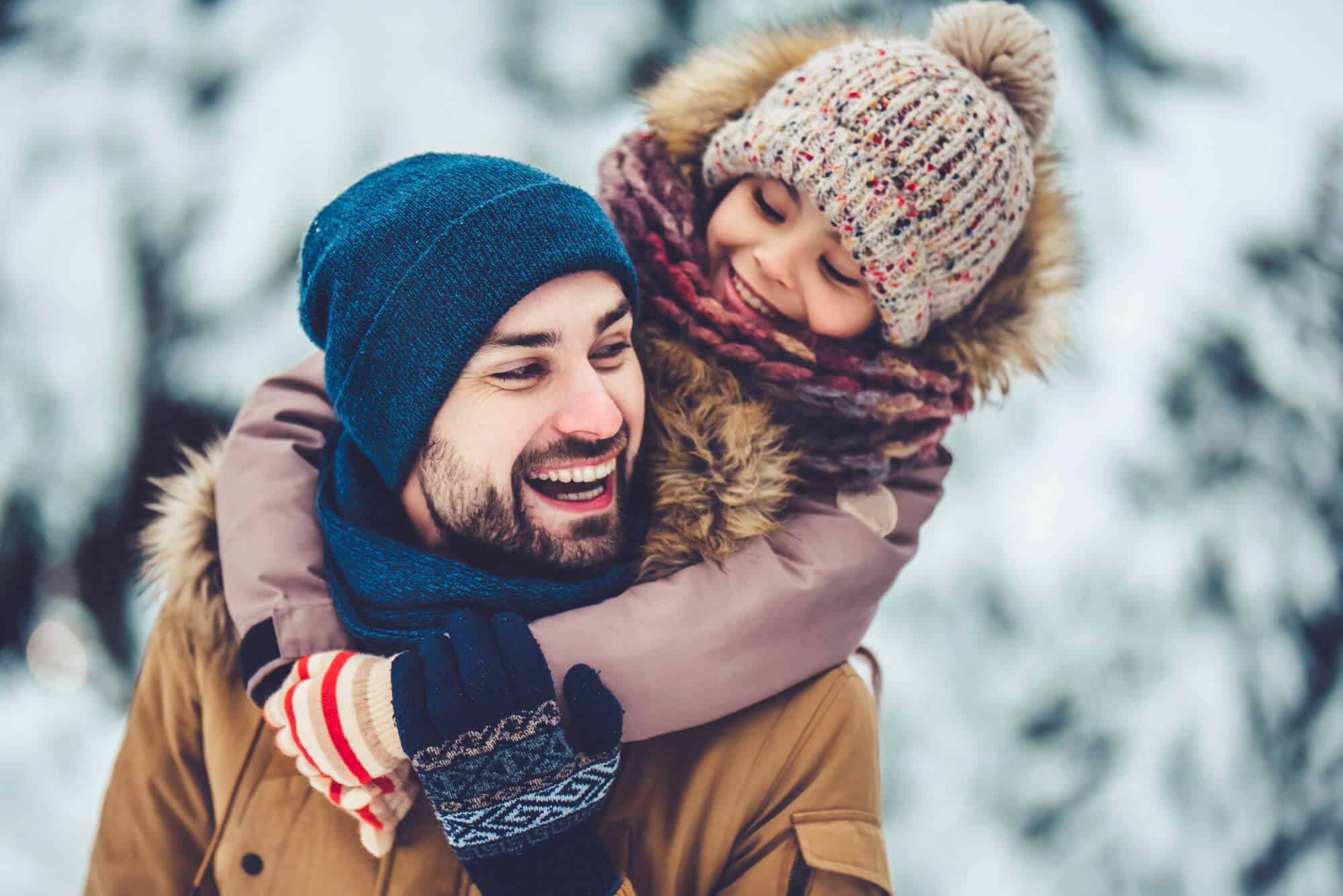 A smiling father wearing a blue beanie and scarf carries his young child in winter clothing on his back, both bundled up warmly in boise id