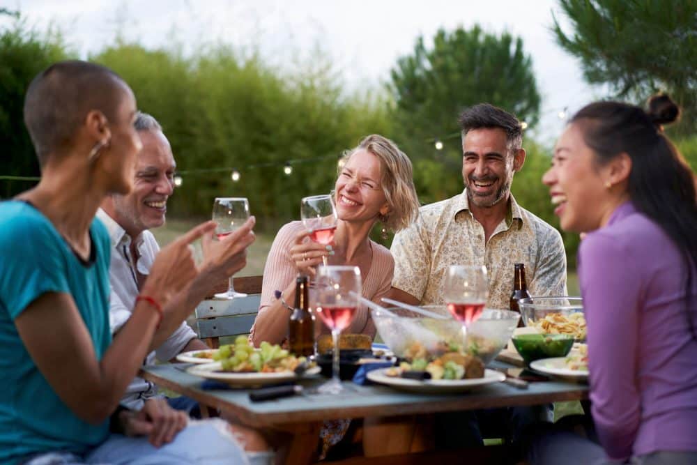 A group of friends sitting at a picnic table outdoors, laughing, enjoying drinks and food, with string lights and greenery in the background in boise id