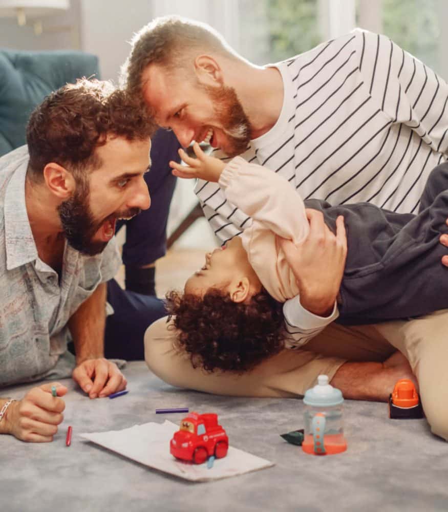 Two fathers laughing and playing with their young child indoors on a rug, surrounded by toys and coloring items in boise id