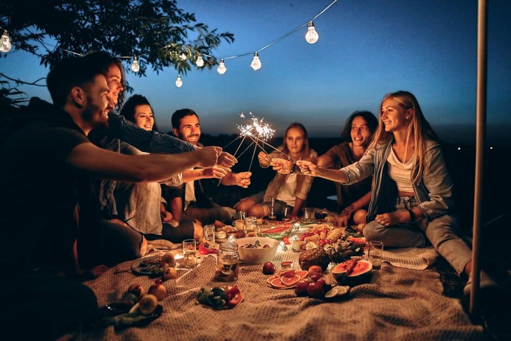 A group of friends enjoys a nighttime picnic under string lights, holding sparklers, surrounded by food and drinks on a blanket in boise id