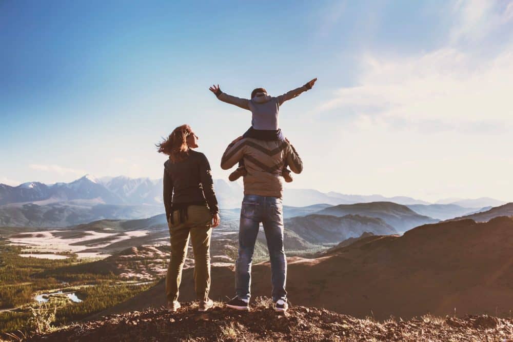 A family stands atop a mountain, with one child sitting on an adult's shoulders, arms outstretched, overlooking a scenic landscape of mountains and valleys in boise id